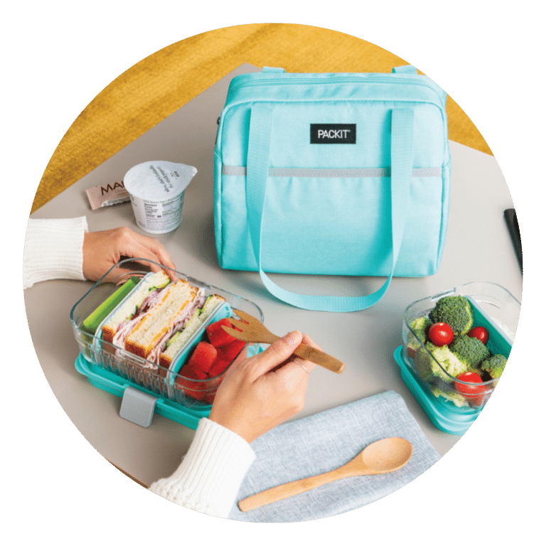 Osoba pakuje turkusową torbę na lunch z napisem PackIt z pudełkiem bento zawierającym kanapkę i truskawki. Na stole znajduje się także inny pojemnik z brokułami i pomidorami, kubek po jogurcie, serwetka i drewniana łyżka.