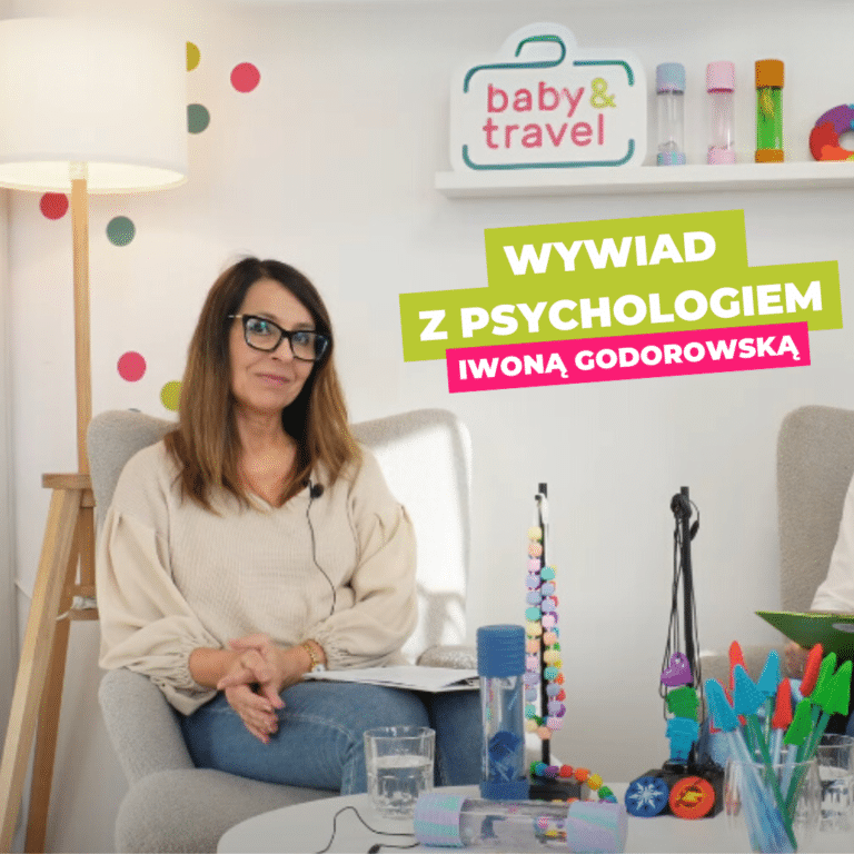 Kobieta w okularach i brązowych włosach siedzi w przytulnym, dobrze oświetlonym pokoju wypełnionym kolorowymi zabawkami i dekoracjami. Ma na sobie beżowy sweter i uśmiecha się do kamery. Za nią widnieje napis "baby & travel", a na neonowym banerze widnieje Ania Kiełbasińska.