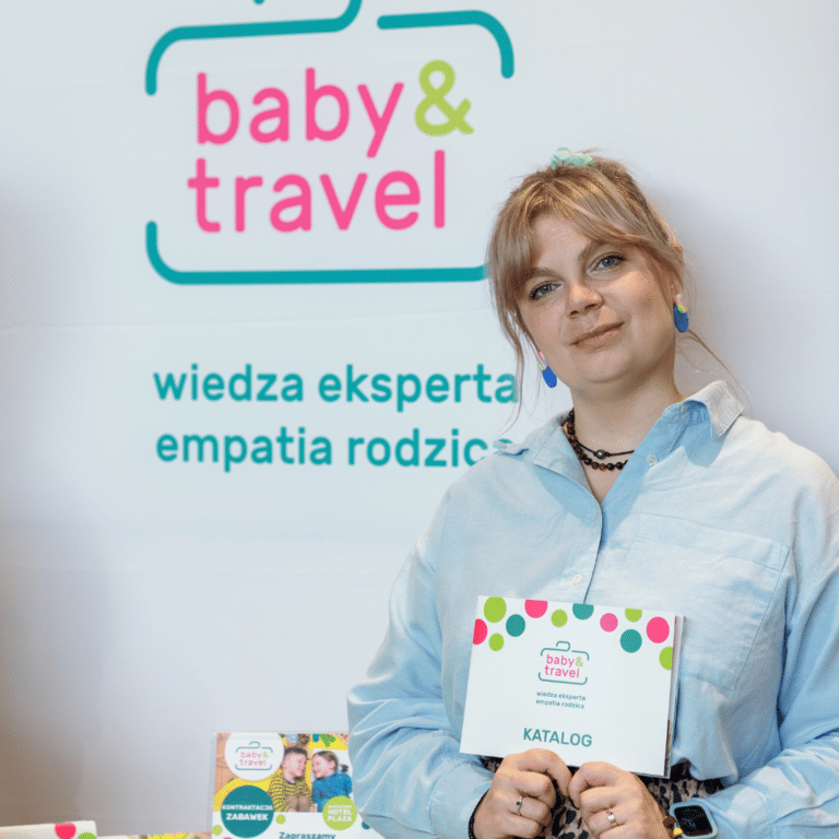 Kobieta z blond włosami spiętymi w kucyk, ubrana w jasnoniebieską koszulę i niebieskie kolczyki, stoi przed banerem baby & travel na targach Klockomania kwiecień 2024. Trzyma kolorowy katalog z tym samym logo; na stole leżą polskie teksty i broszury.